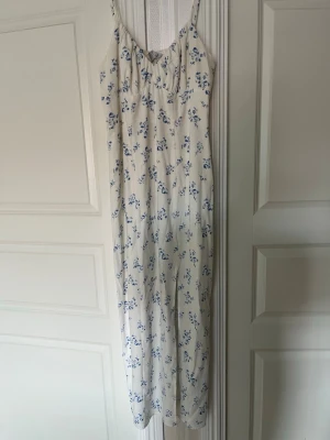 Vit långklänning med blå blommor - Söt långklänning i vit med blått blommönster och smala axelband. Klänningen har en rak passform och är ärmlös, perfekt för varma dagar. Lätt och luftig känsla, passar till sommarens alla tillfällen.