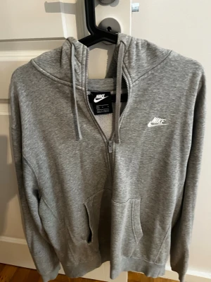 Grå zip hoodie från Nike - Snygg grå hoodie från Nike med dragkedja framtill och klassisk logga broderad på bröstet. Hoodien har huva med dragsko och två stora fickor framtill. Tillverkad i mjukt bomullsmaterial som är skönt att ha på sig. Perfekt för en avslappnad och sportig stil.