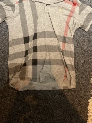Grå rutig t-shirt från Burberry - Säljer en grå t-shirt från Burberry med klassiskt rutigt mönster i svart, rött och vitt. T-shirten har korta ärmar och en liten broderad Burberry-logga på bröstet. Tillverkad i mjuk bomull för skön känsla.