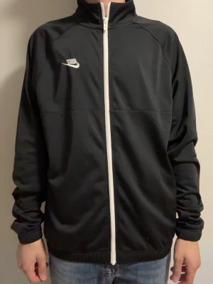 Svart kofta från Nike - Svart kofta från Nike med vit dragkedja och logga på bröstet. Jackan har hög krage, lång ärm och en sportig look. Perfekt för dig som gillar stilren och enkel design.