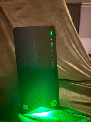 HP stationär gaming dator svart - Säljer en svart stationär HP gaming dator med stilren design och grön LED-belysning framtill. Framsidan har flera USB-portar och hörlursuttag. Chassit är gjort av plast och metall, och har HP-loggan längst ner. Perfekt för gaming eller studier. Den är ny damsugen och återställd som ny. Den har en RTX 3060 TI, intel 5 11400f 32 gb DDR4 kingston fury ram med 512 GB ssd med en 2 terabyte hårddisk 