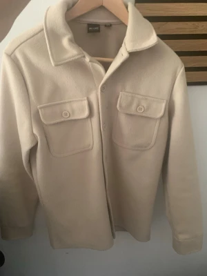 Beige overshirt från Only & Sons - Stilren beige overshirt från Only & Sons med två stora bröstfickor med lock och knappar. Skjortjackan har klassisk krage, raka ärmar och är gjord i ett mjukt, fleece-liknande material. Perfekt lager på lager-plagg för en avslappnad look.