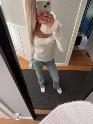 Vit ribbad topp med volang - Supersöt vit ribbad topp med lång ärm och bred volang runt den fyrkantiga ringningen. Toppen har en figurnära passform och är tillverkad i ett mjukt, stretchigt material som känns bekvämt mot huden. Perfekt att styla med jeans eller kjol för en trendig look.använd en gång!!