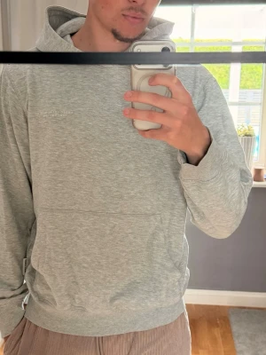 Ljusgrå hoodie  - Säljer en ljusgrå hoodie från J&J med diskret logga framtill och stor tryckt text på ryggen. 