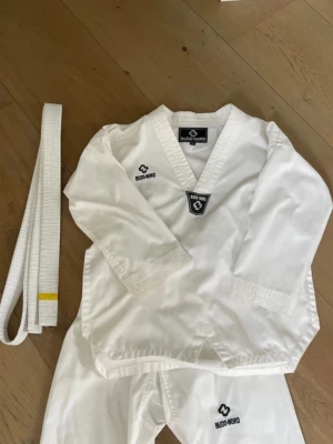 Barn taekwondodräkt från Budo-Nord - Vit taekwondodräkt från Budo-Nord i bomull. Setet innehåller långärmad överdel med V-ringning, byxor med resår i midjan och ett vitt bälte med gul markering. Perfekt för dig som tränar kampsport och vill ha en klassisk look.
