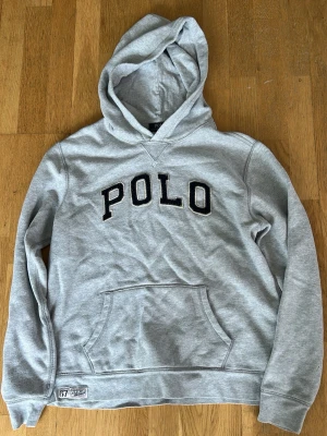 Grå hoodie från Polo Ralph Lauren - Snygg grå hoodie från Polo Ralph Lauren med svart broderad POLO-text på bröstet. Klassisk känguruficka framtill och huva. Mjuk bomullsblandning som är perfekt för en avslappnad stil.