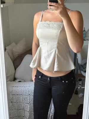 Beige linnetopp med spetsdetalj - Supersöt beige linnetopp med smala axelband och vacker spets upptill. Toppen har en lätt utsvängd passform och är perfekt för en trendig och avslappnad look. Spetsen har blommigt mönster som ger en romantisk touch.