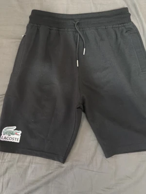 Svarta shorts från Lacoste med snörning - Snygga svarta shorts från Lacoste med klassisk krokodillogo på vänster ben. Shortsen har snörning i midjan och sidofickor. Perfekta för en avslappnad och sportig stil.
