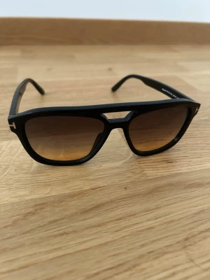 Svarta Tom Ford solglasögon - Snygga svarta solglasögon från Tom Ford med markerad båge och bruna tonade glas. Klassisk fyrkantig form med diskret logga på sidan. Ramen är i plast och ger en modern och stilren look.