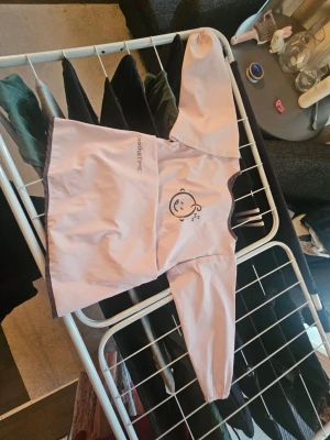 Rosa haklapp med ärmar - Söt rosa haklapp med långa ärmar och brunt kantband. Framtill finns ett gulligt tryck av ett sovande ansikte och texten 'SNACK&TIME'. Perfekt för småbarn vid måltider eller pyssel. Tillverkad i lätt avtorkbart material.