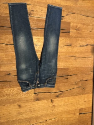 Blå jeans med slitningar och knappar - Säljer ett par blå jeans med slitningar och knappar i gylfen. Jeansen har klassisk femficksdesign och kontrastsömmar. Färgen är mörkblå med ljusa partier och detaljer som ger en cool, använd look. Dom är sjukt stora för dess storlek men är ett sjukt snyggt plagg 