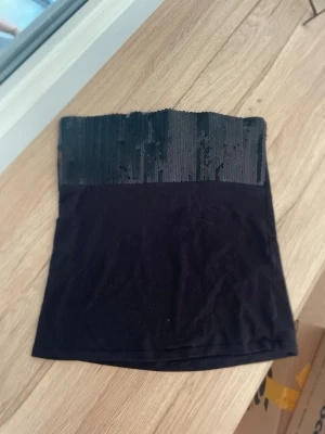 Svart bandeau tubtopp med paljetter - Snygg svart bandeau tubtopp med glittriga paljetter upptill. Toppen är axelbandslös och har en enkel, tight passform som sitter på plats. Perfekt för dig som vill ha en stilren men ändå festlig look.