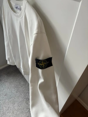 Vit sweatshirt från Stone Island - Vit sweatshirt från Stone Island med klassisk rund hals och ikonisk svart patch med logga på vänster ärm. Tröjan har en enkel och stilren design, ribbade muddar vid ärmslut och nederkant. Perfekt för dig som gillar streetwear och vill ha en clean look.
