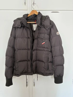 Svart dunjacka från Moncler - Snygg svart dunjacka från Moncler med avtagbar huva, ribbade muddar och klassisk logga på ärmen. Jackan har två stora fickor framtill och stängs med dragkedja och knappar. Perfekt för kalla vinterdagar och riktigt stilren.