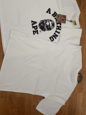 Vit A Bathing Ape t-shirt - Säljer en vit t-shirt från A Bathing Ape med det klassiska BAPE-logotrycket i svart och grått kamouflagemönster på bröstet. T-shirten har korta ärmar och rund hals. Perfekt för dig som gillar streetwear och vill ha något stilrent men ändå ikoniskt.