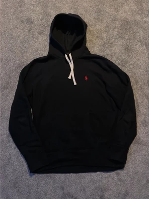 Svart hoodie från Polo Ralph Lauren - Svart hoodie från Polo Ralph Lauren med vit dragsko och den klassiska röda logotypen broderad på bröstet. Hoodien har känguruficka framtill och är tillverkad i mjukt bomullsmaterial. Perfekt för en avslappnad och stilren look.