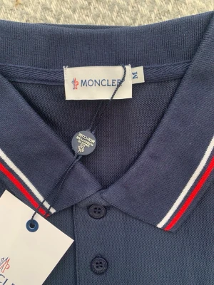 Mörkblå Moncler pikéskjorta - Snygg mörkblå pikéskjorta från Moncler med klassisk krage och knappar framtill. Kragen har röda och vita ränder som ger en sportig touch. Moncler-logga broderad på bröstet. Perfekt för en stilren och avslappnad look.