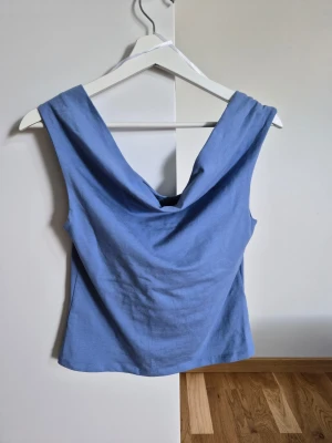 Ljusblå ärmlös topp med draperad urringning - Snygg ljusblå topp i mjuk bomull med draperad urringning både fram och bak. Modellen är ärmlös och har en avslappnad passform som ger en cool och trendig look. Perfekt att matcha med jeans eller kjol för en stilren outfit.