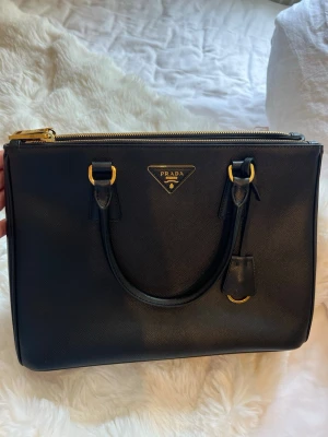 Svart handväska från Prada - Snygg svart handväska från Prada i klassisk boxig form med guldfärgade metalldetaljer och dubbla handtag. Väskan är tillverkad i strukturerat läder och har Pradas ikoniska logga framtill. Medföljer original dustbag. Finns även en orginal axelband rem om man även vill ha väska över axeln. Även remen i orginal dusty bag. Äkthetsbevis medföljer och kvittot finns att få se om man vill 