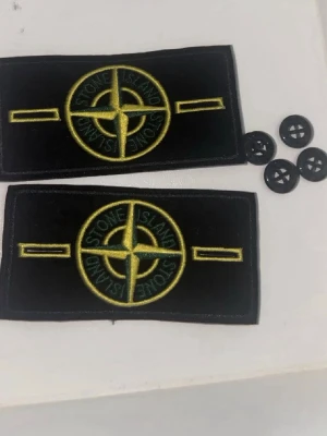 Stone Island tygmärken med knappar - Två svarta tygmärken från Stone Island med det klassiska gula och gröna kompasslogotypen. Märkena är rektangulära och kommer med fyra svarta knappar för fastsättning. Perfekt för att sätta på jackor, tröjor eller väskor.