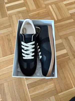 Svarta maison margiela gats - Stilrena svarta sneakers med beige sula och vita snören. Ovandelen är i en mix av mocka och slätt läder, vilket ger en snygg kontrast. Insidan är ljus och skön. Perfekta för dig som gillar klassisk men modern stil.