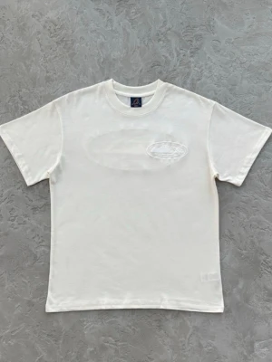 Corteiz | Vit t-shirt - Vit t-shirt från Corteiz med ett subtilt, glansigt tryck i form av en oval och motiv på bröstet. Klassisk passform med rund hals och korta ärmar. Perfekt för en clean och stilren look.