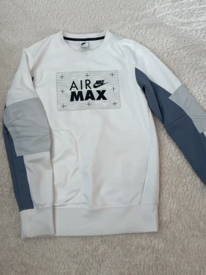 Nike Air Max vit och grå hoodie - Nike Air Max hoodie i vitt med grå detaljer på ärmarna och stor svart logga framtill. Tröjan har rund halsringning och är tillverkad i mjukt material som känns skönt mot huden. Perfekt för en sportig och avslappnad stil.