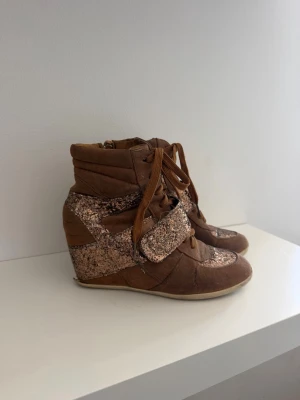 Bruna sneakers med kilklack och glitter - Unika bruna sneakers med kilklack och glittriga detaljer i rosa och guld. Skorna har snörning och kardborreband över vristen för extra stil och passform. Materialet är en mix av mocka och textil med glitterpaneler som ger en trendig look.