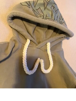 Syna world hoodie - Snygg beige hoodie med stora vita dragsnören och tryck på huvan. Hoodien har en klassisk passform och är tillverkad i mjukt bomullsmaterial. Perfekt för en avslappnad och trendig look.