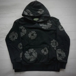 Denim Tears | Svart hoodie - Cool svart hoodie från Denim Tears med unika gråa blommönster över hela tröjan. Klassisk huva och känguruficka framtill. Perfekt för dig som gillar streetwear och vill sticka ut med en kreativ design.