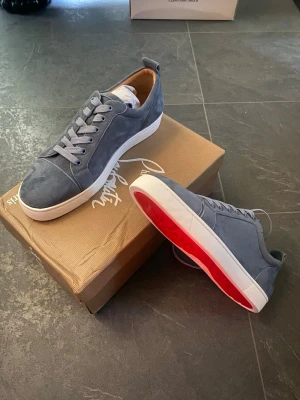 Gråa Louboutin sneakers i mocka - Snygga gråa sneakers från Christian Louboutin i mjuk mocka med klassisk röd sula och vita kanter. Skorna har rund tå, gråa skosnören och diskret branding på insidan. Perfekta för dig som vill ha en clean och exklusiv look.