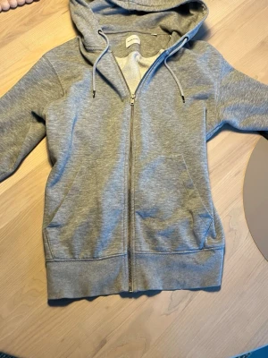 Grå zip hoodie från Jack & Jones - Säljer en klassisk grå zip hoodie från Jack & Jones. Hoodien har dragkedja framtill, två fickor och justerbar huva med snören. Tillverkad i mjukt sweatshirtmaterial som är borstat på insidan för extra komfort. Perfekt för en avslappnad och stilren look.