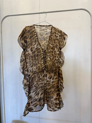 Leopardmönstrad strandtunika med snörning - Säljer en transparent strandtunika med leopardmönster och djup V-ringning med snörning framtill. Tunikan har guldfärgade nitar längs ringningen och är tillverkad i ett lätt, luftigt material. Perfekt att slänga över bikinin på stranden.
