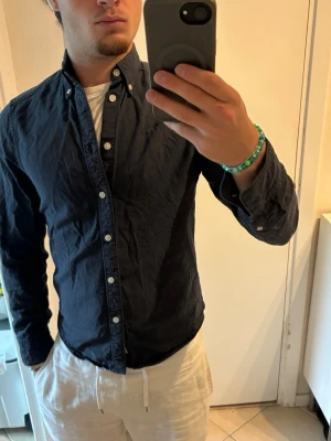Mörkblå skjorta från east west  - Klassisk mörkblå skjorta från east west med vita knappar och button down-krage. Skjortan har lång ärm och är tillverkad i bomull, vilket gör den både bekväm och enkel att styla till olika outfits. Tröjan är urtvättad, se egentlig färg på bild 3. 