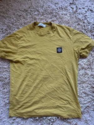 Gul t-shirt från Stone Island - Säljer en gul t-shirt från Stone Island med klassisk rund hals och korta ärmar. T-shirten har en svart patch med logga på bröstet och är tillverkad i mjuk bomull. Perfekt för dig som gillar streetwear och enkel stil. Liten i storlek, väldigt bra skick. Är äkta men har klippt bort lapparna då de var obehagliga. Kostar 1800 ny
