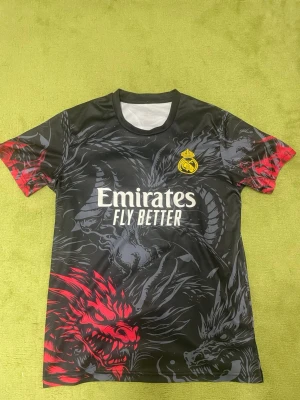 Real Madrid draktröja svart/röd - Cool svart sporttröja från Real Madrid med drakmotiv i grått och rött. Tröjan har korta ärmar, rund hals och klubbens gula logga på bröstet. Tryck med 'Emirates Fly Better' på framsidan. Perfekt för fotboll eller träning.