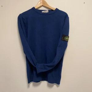 Blå Stone Island stickad tröja - Säljer vidare en blå stickad tröja från Stone Island med klassisk patch på vänster ärm. Tröjan har rund halsringning, ribbade muddar och lång ärm. Perfekt för dig som gillar streetwear och vill ha något stilrent men ändå med tydlig logga.