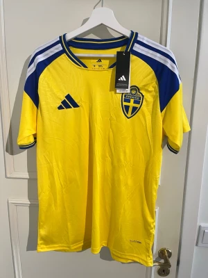 Sverige matchtröja Adidas gul/blå - Säljer en officiell Sverige matchtröja från Adidas i gult med blå detaljer och vita ränder på axlarna. Tröjan har Sveriges landslagsemblem på bröstet och 'Sverige' tryckt i nacken. Tillverkad i lätt och ventilerande Climalite-material.