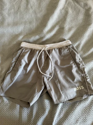 Gråa  badbyxor från BOSS - Snygga gråa badbyxor från BOSS med vit dragsko i midjan och diskret logga på vänster ben. Klassisk modell med sidofickor och elastisk midja. Tillverkade i snabbtorkande material, perfekta för stranden eller poolen.