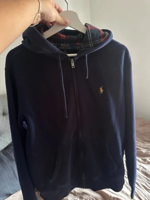Mörkblå hoodie från Polo Ralph Lauren - Mörkblå hoodie med dragkedja från Polo Ralph Lauren. Klassisk gul logga på bröstet och huva med rödgrönt rutigt foder. Nypris runt 1500