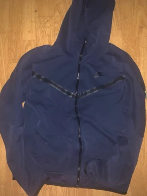 Mörkblå Nike hoodie med dragkedja - Snygg mörkblå hoodie från Nike med hel dragkedja framtill och svarta detaljer över bröstet. Klassisk huva och diskret Nike-logga på vänster sida. Tillverkad i mjukt material som känns skönt mot huden. Perfekt för en sportig och avslappnad stil.