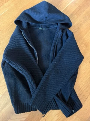 Svart stickad hoodie med dragkedja - Mysig svart stickad hoodie från bik bok med dragkedja framtill och stor huva. Tröjan har ribbad struktur och långa ärmar, perfekt för en avslappnad och cool stil. Passar dig som gillar streetwear och vill ha något bekvämt men ändå snyggt.