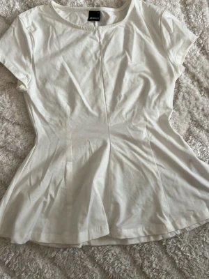 Vit topp från Gina Tricot - Säljer en enkel och stilren vit topp från Gina Tricot. Modellen har korta ärmar, rund halsringning och lätt insvängd midja för en feminin siluett. Perfekt att matcha med jeans eller kjol för en clean look.