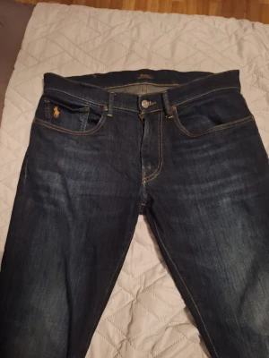 Mörkblå jeans från Polo Ralph Lauren - Säljer ett par mörkblå jeans från Polo Ralph Lauren med klassisk femficksmodell och gul kontrastsöm. Jeansen har en diskret logga på myntfickan och är tillverkade i denim med lätt tvättad look. Perfekta för en stilren och avslappnad outfit.