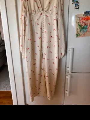 Vit blommig långklänning med knappar adoore - Söt långklänning i vit med små röda blommor, v-ringad och med knappar framtill. Klänningen har långa ärmar och är fodrad. Perfekt för sommarens alla tillfällen!