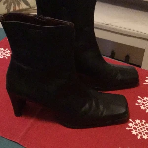 Svarta skinnstövletter med klack 7cm - Snygga svarta stövletter i skinn med dragkedja på sidan och en elegant klack 7 cm. Perfekta för att ge en stilren touch till din outfit. De har en spetsig tå och är tillverkade i ett glansigt material. Made in Italy. Tamaris.