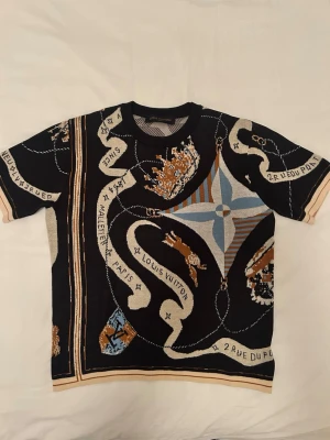 Louis Vuitton mönstrad t-shirt - Unik svart t-shirt från Louis Vuitton med detaljerat mönster i beige, blått och brunt. T-shirten har korta ärmar och rund hals. Materialet känns som bomull och trycket har inslag av text och grafiska former som ger en exklusiv vibe.