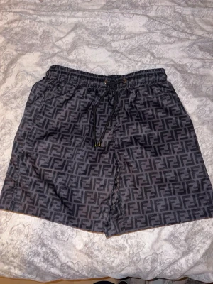 Svarta Fendi shorts med logomönster - Snygga svarta shorts från Fendi med hela shortsen täckta av det klassiska FF-logomönstret. De har elastisk midja med snörning och är tillverkade i ett glansigt syntetmaterial. Perfekta för dig som vill sticka ut med en lyxig streetstyle.