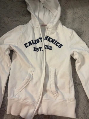 Vit zip up hoodie! - Vit hoodie med dragkedja och blått tryck 'CALISTHENICS EST. 2002' på bröstet.  Använd 2 gånger och är för liten för mig! 