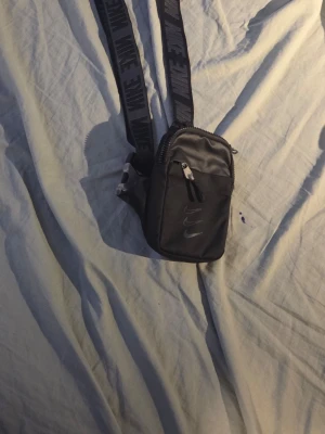 Svart axelväska från Nike - Snygg svart axelväska från Nike med justerbar rem där det står 'NIKE' i grått. Väskan har två dragkedjefack och en liten Nike-logga framtill. Perfekt storlek för mobil, plånbok och nycklar. Materialet är slitstark nylon.